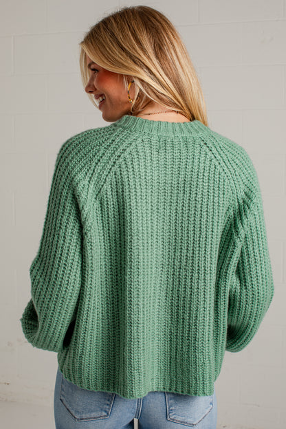 A woman wearing a sage green chunky knit sweater, shown from the back against a white brick wall.