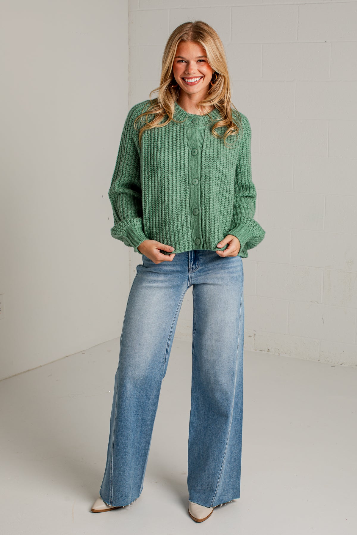 A woman modeling a sage green cable-knit cropped cardigan with button-front closure and wide-leg light-wash blue jeans, standing against a light gray studio wall.