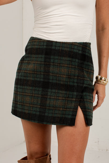 A close-up of a model wearing a dark green and black plaid mini skirt with a side slit, styled with a white top and brown boots.