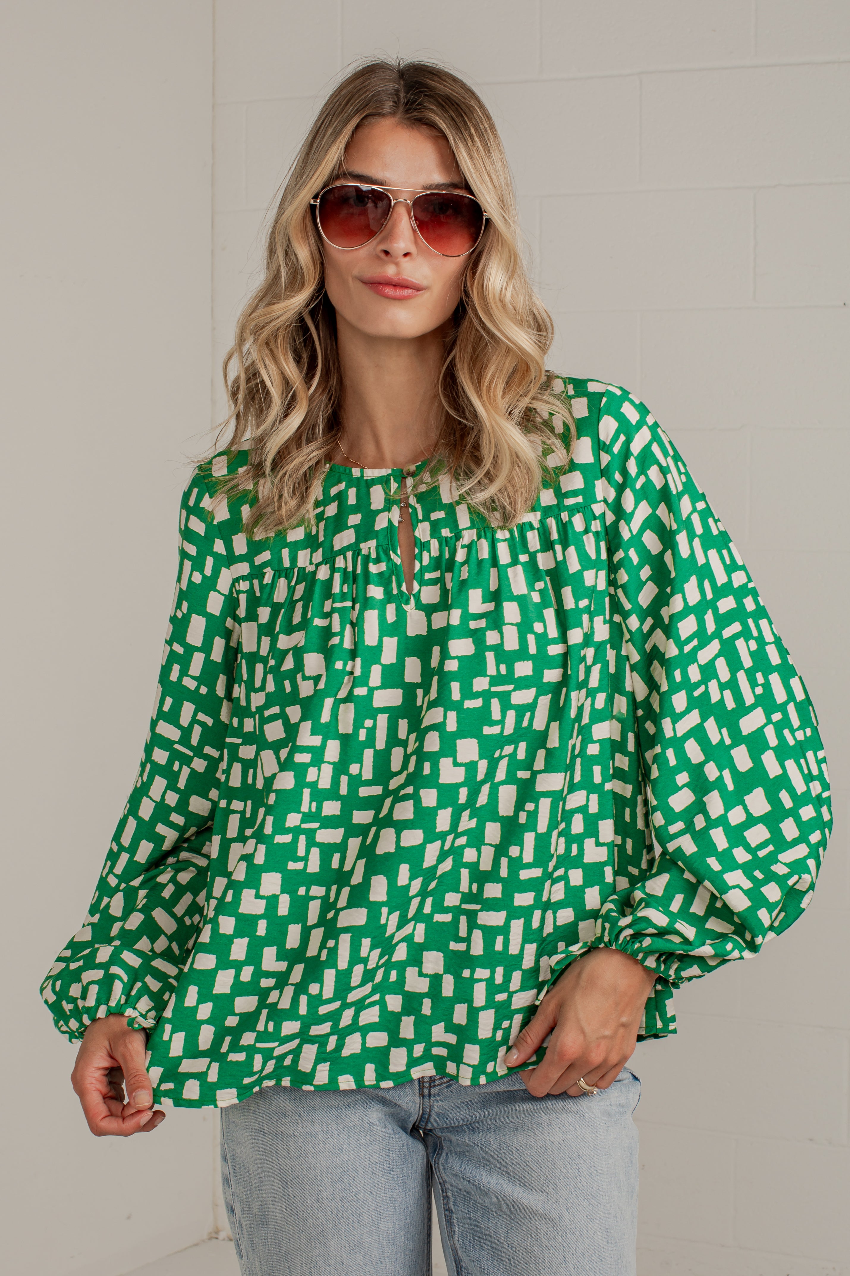 A woman wearing a green and white geometric patterned blouse with balloon sleeves and a keyhole neckline, paired with light blue jeans and sunglasses, standing against a light gray brick wall.
