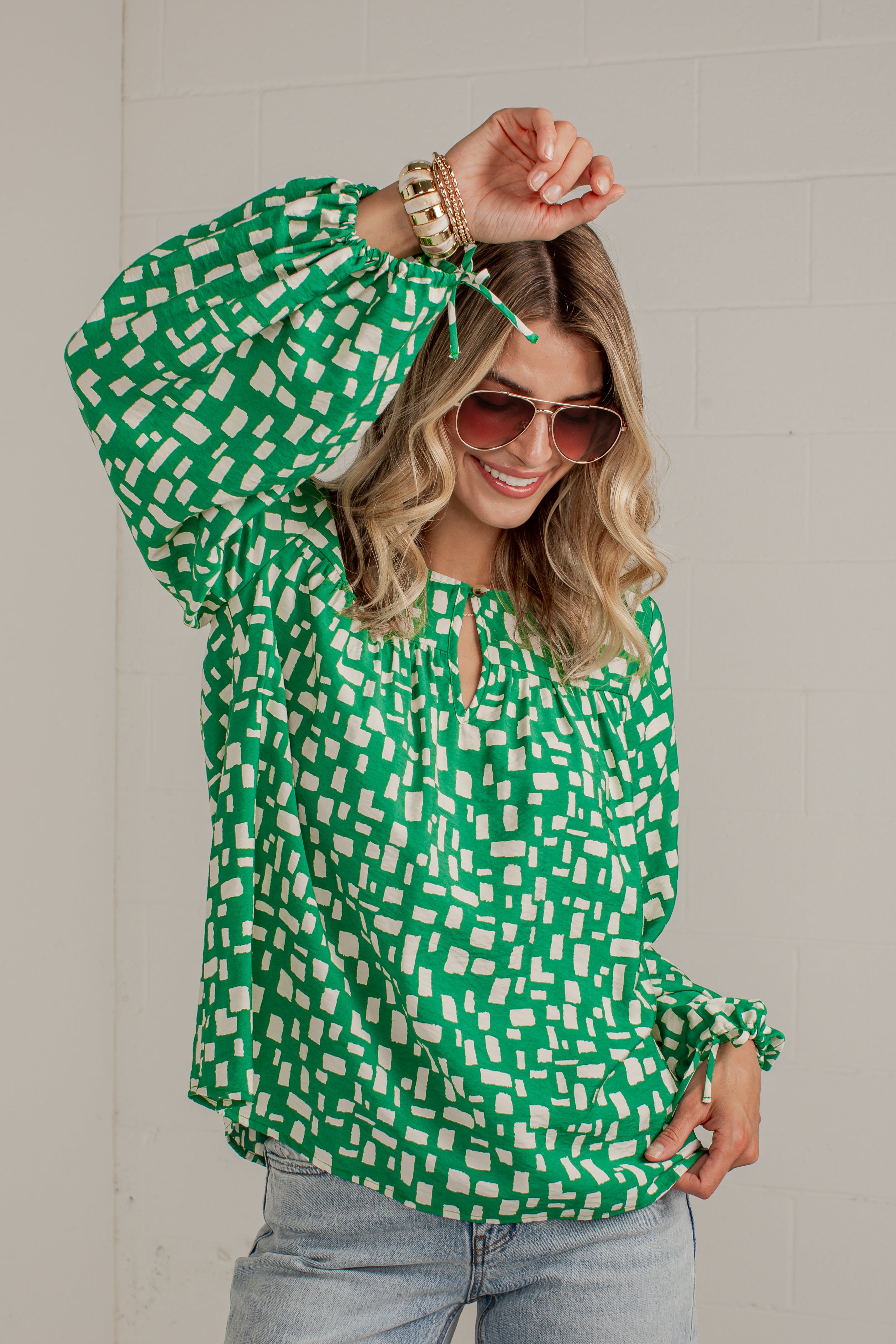 A woman wearing a green and white geometric patterned blouse with long balloon sleeves and a keyhole neckline, paired with light blue jeans and sunglasses, posing against a light gray brick wall.