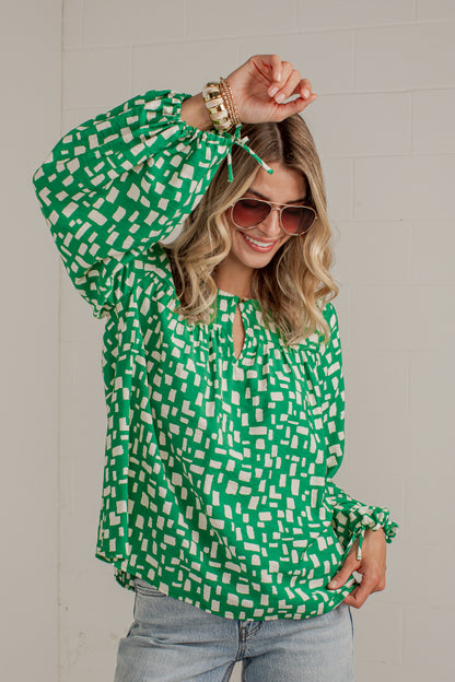 A woman wearing a green and white geometric patterned blouse with long balloon sleeves and a keyhole neckline, paired with light blue jeans and sunglasses, posing against a light gray brick wall.