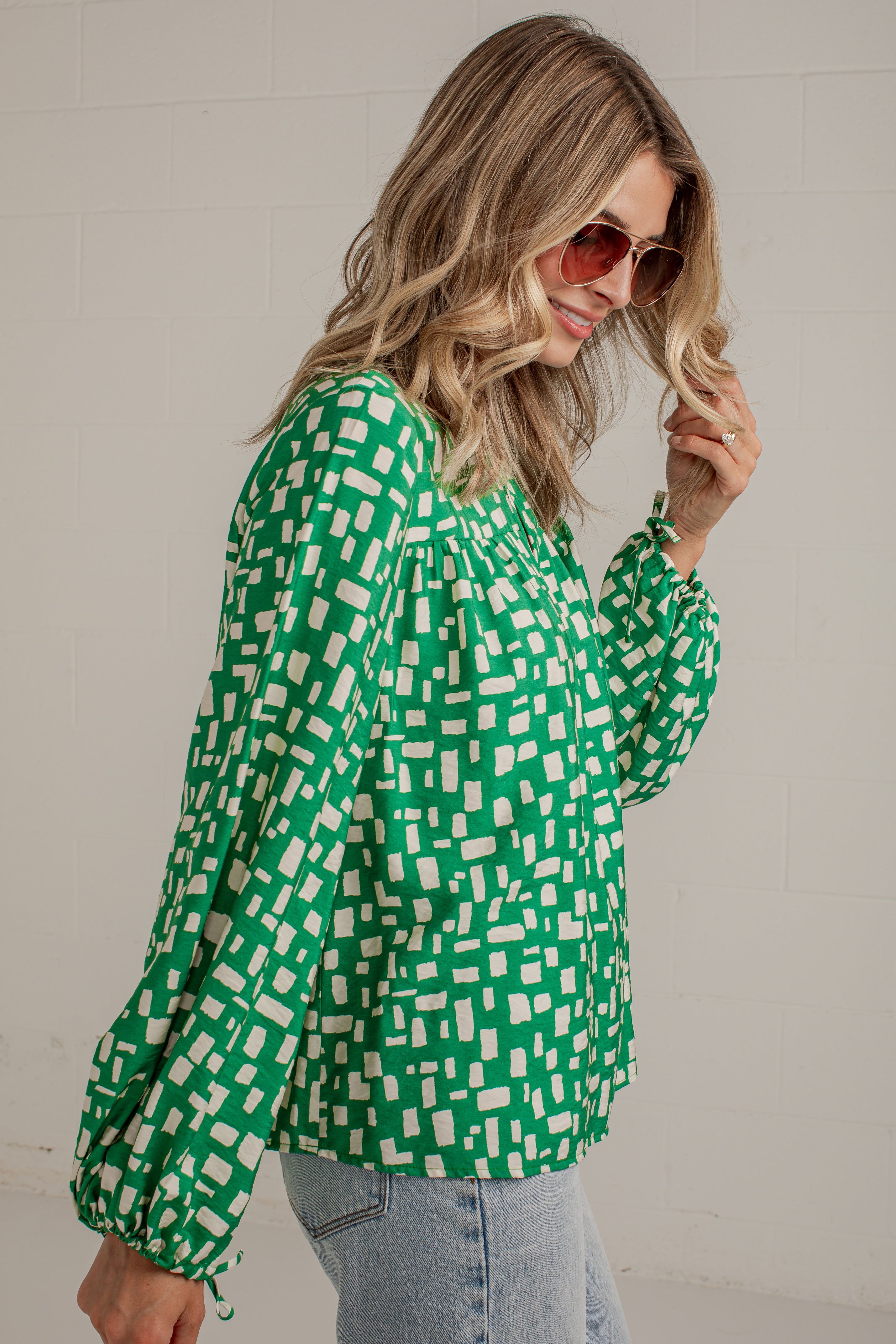 A woman wearing a green and white geometric patterned long-sleeve blouse with light blue jeans, posing against a white brick wall.
