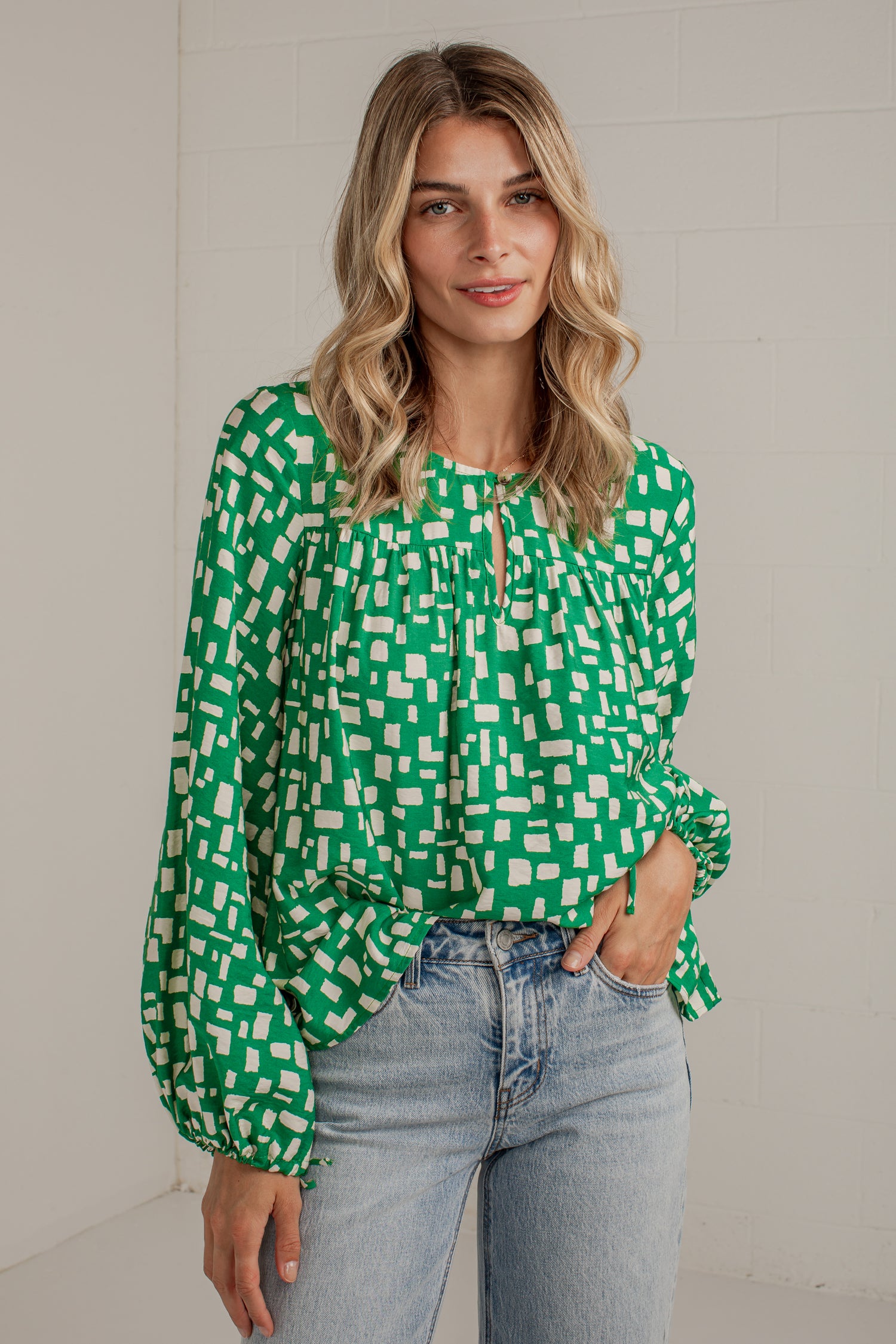 A woman wearing a green and white geometric patterned blouse with long balloon sleeves and light-wash blue jeans, standing against a white brick wall.