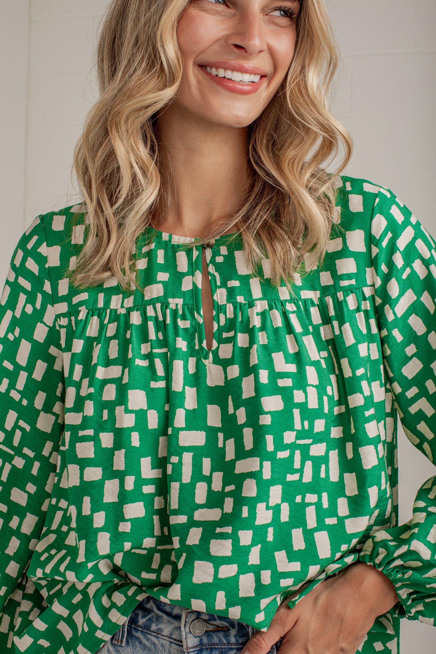 A woman wearing a green long-sleeve blouse with a white geometric block pattern and a keyhole neckline, paired with blue jeans.