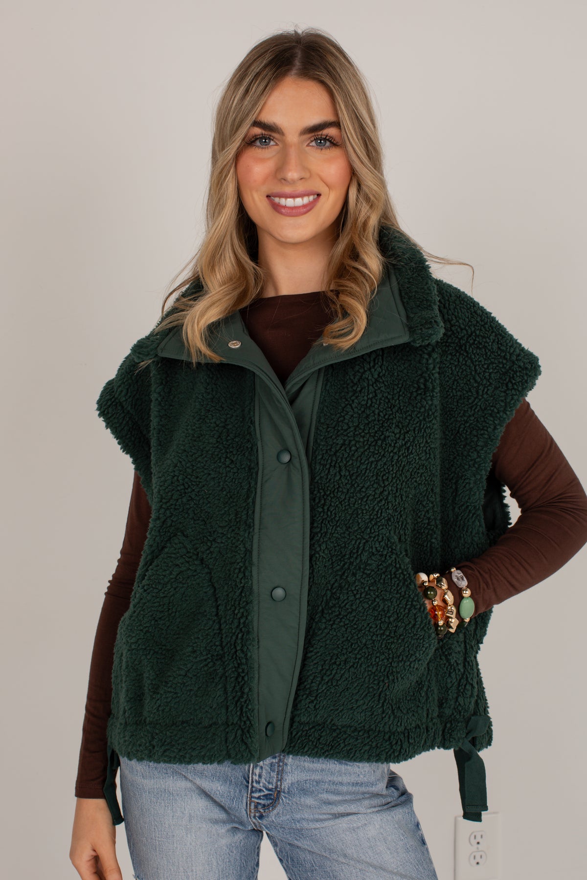 A woman wearing a dark forest green sherpa fleece vest over a brown long-sleeve top and light blue jeans, standing against a plain light gray studio background.