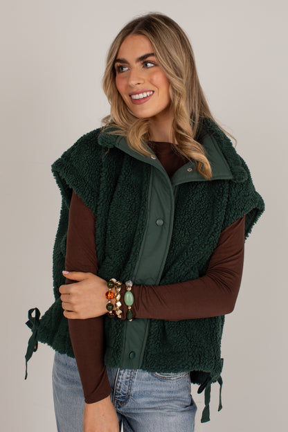 A woman wearing a dark forest green sherpa fleece vest over a brown long-sleeve top and blue jeans, posed against a solid light gray studio background.