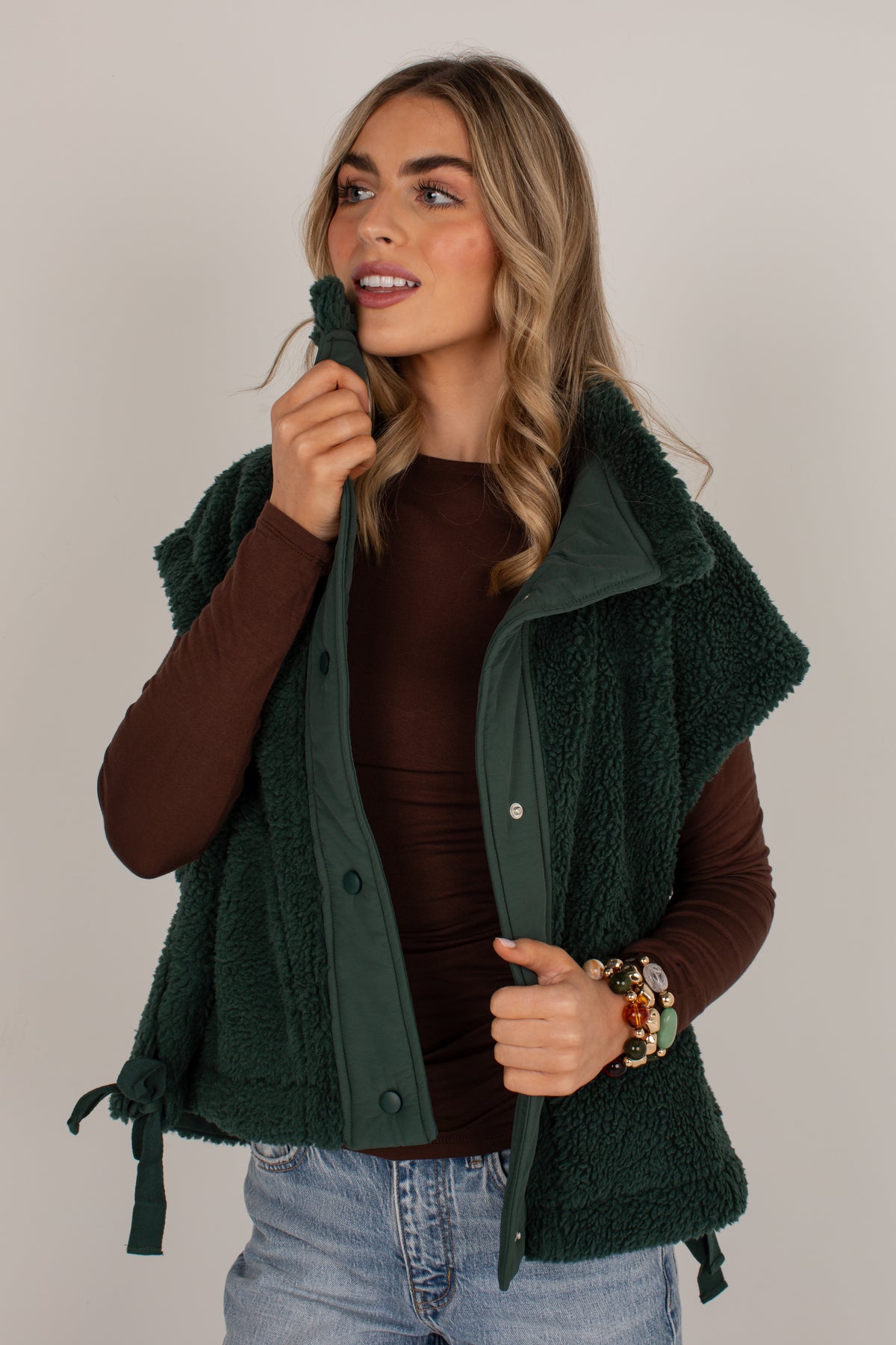 A woman wearing a dark forest green sherpa fleece vest over a brown long-sleeve top and light blue jeans, posing against a neutral light gray background.