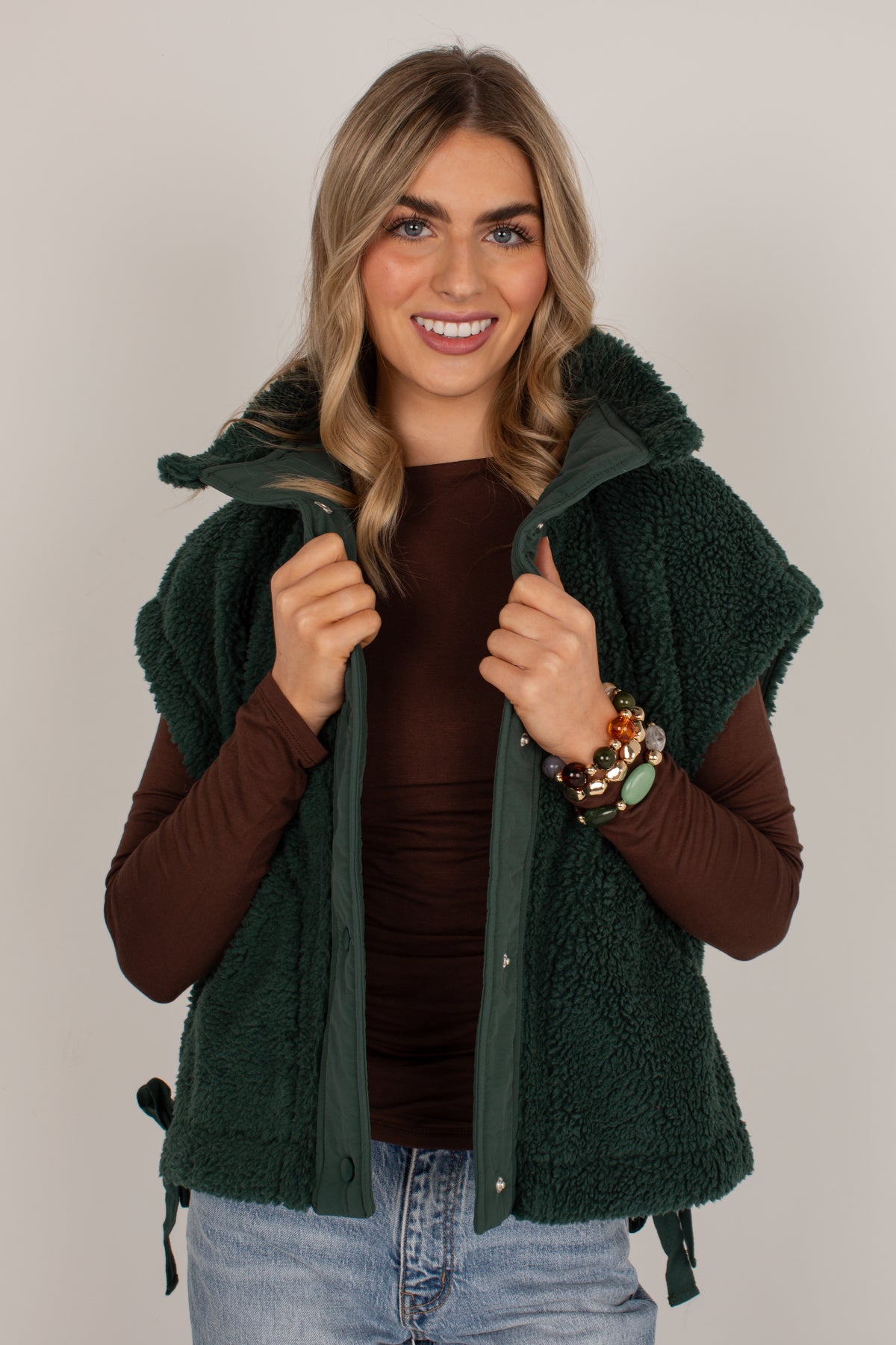 A smiling blonde woman wearing a dark forest green sherpa fleece vest over a brown long-sleeve top and light blue jeans, standing against a neutral light gray studio background.