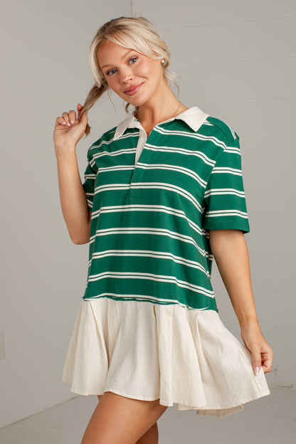 A woman modeling a green and white striped short-sleeve polo shirt with a cream pleated skirt, standing against a light gray wall.