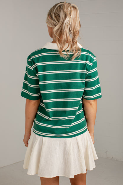 A woman viewed from behind wearing a green short-sleeve polo shirt with thin white horizontal stripes and a white pleated skirt, standing against a light gray wall.