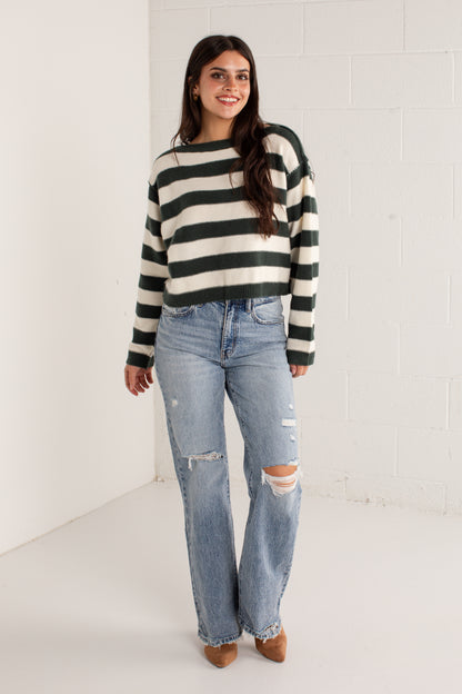A woman modeling a dark green and cream striped knit sweater paired with light-wash distressed jeans and brown pointed-toe heels, standing against a white brick wall.