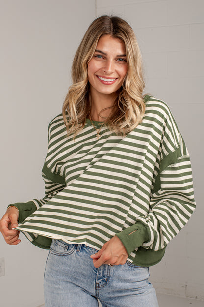 A woman wearing a green and cream striped long-sleeve top with solid green cuffs and hem, paired with light blue jeans, standing against a light gray wall.