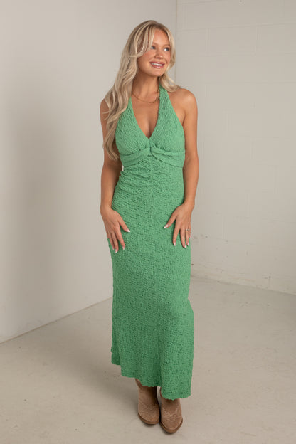 A woman modeling a green textured halter-neck maxi dress in a minimalist studio setting with off-white walls and beige flooring.