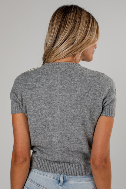 A woman viewed from behind wearing a short-sleeve gray knit sweater and light blue jeans against a plain light gray studio background.