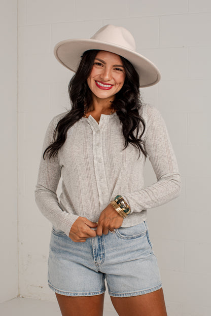 A woman wearing a light gray ribbed knit long-sleeve button-up shirt, light-wash denim shorts, a beige wide-brim hat, and gold bracelets, standing against a white brick wall.