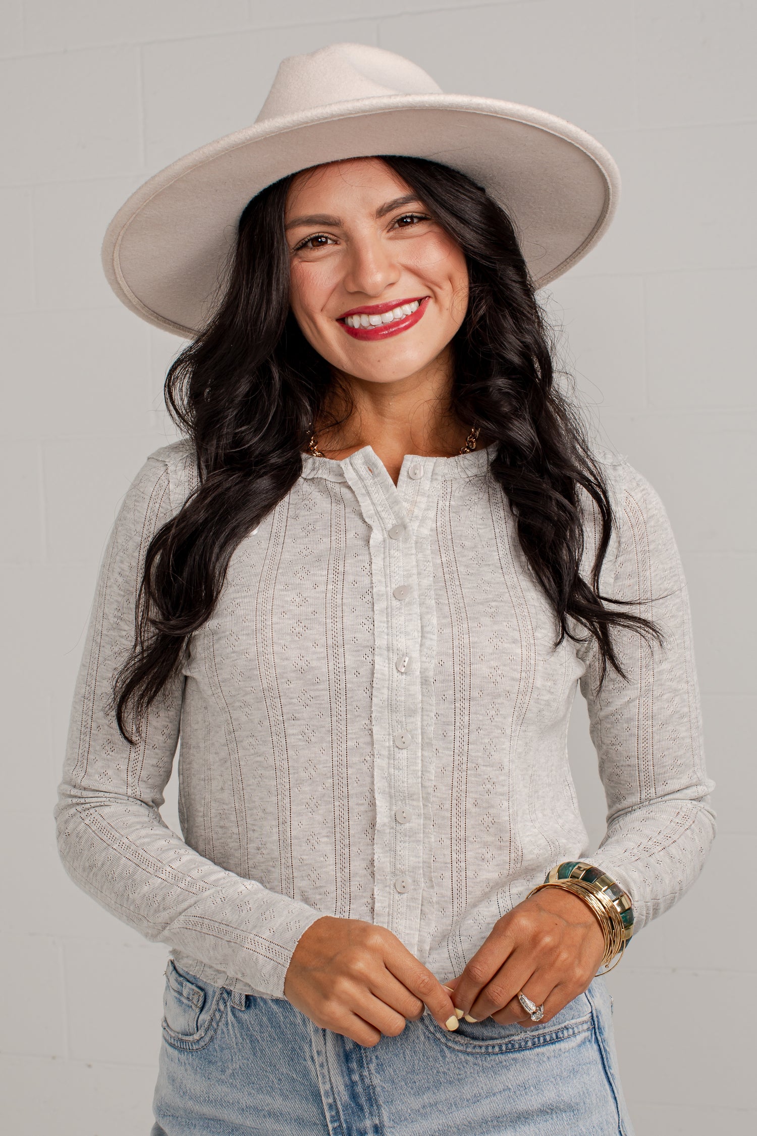 A smiling woman wearing a light beige wide-brim hat and a light gray long-sleeve button-up shirt, paired with blue jeans, against a light gray brick wall background.