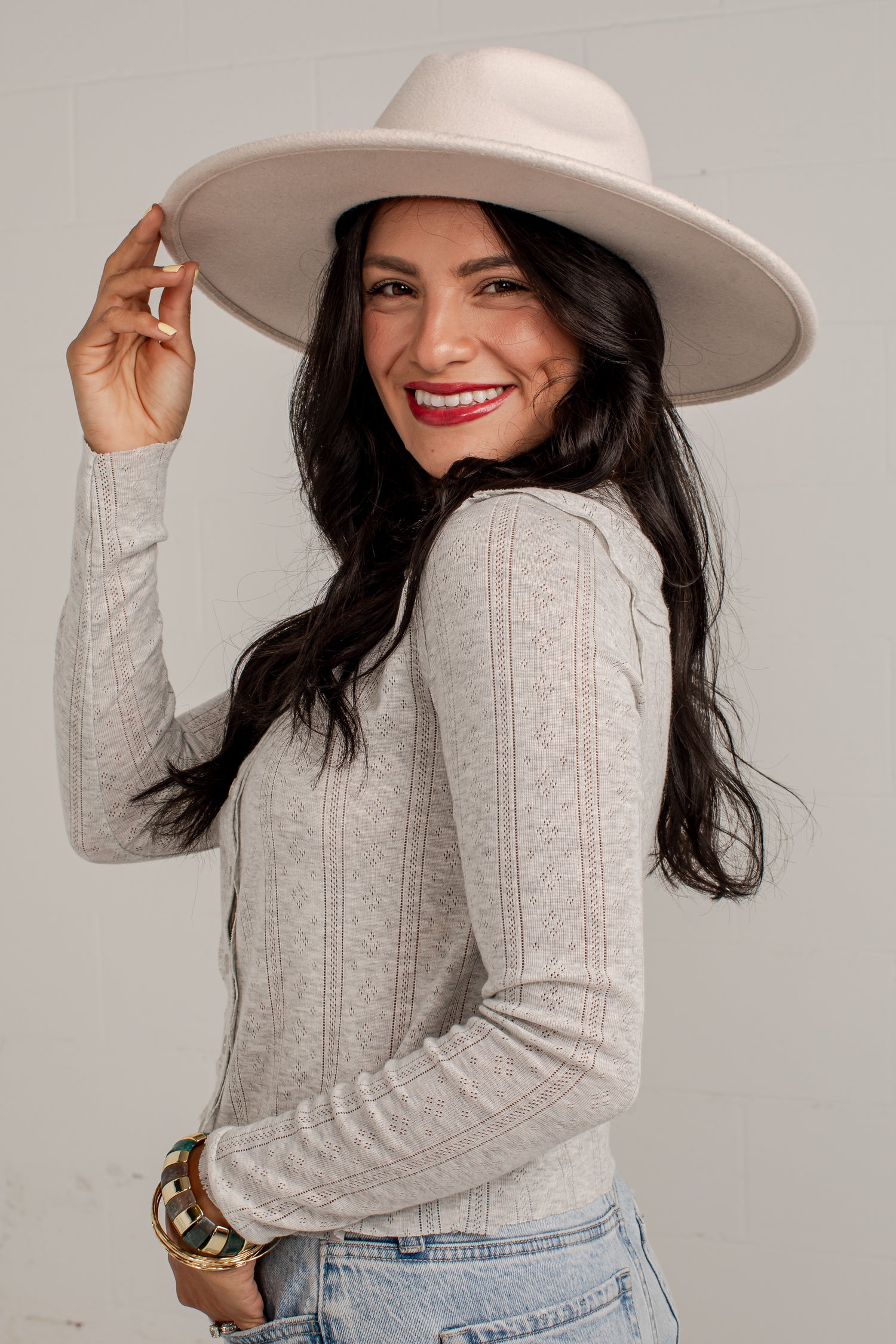 A woman wearing a light beige wide-brim fedora hat and a light gray ribbed knit long-sleeve top, paired with light-wash blue jeans. She is smiling and looking over her shoulder against a light gray background.