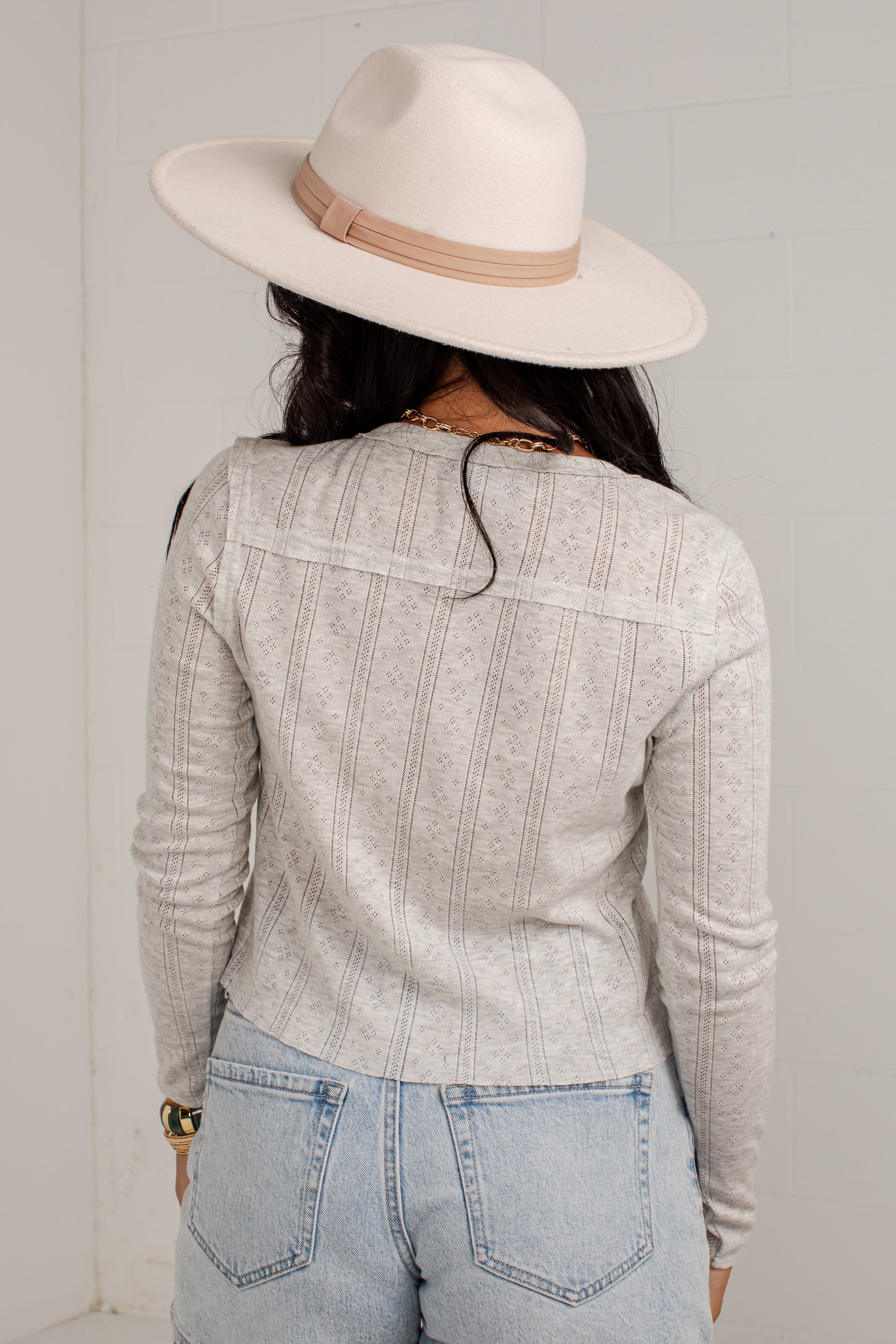 A woman shown from behind wearing a light gray ribbed knit long-sleeve top, light blue jeans, and a cream wide-brim fedora hat with a tan band.