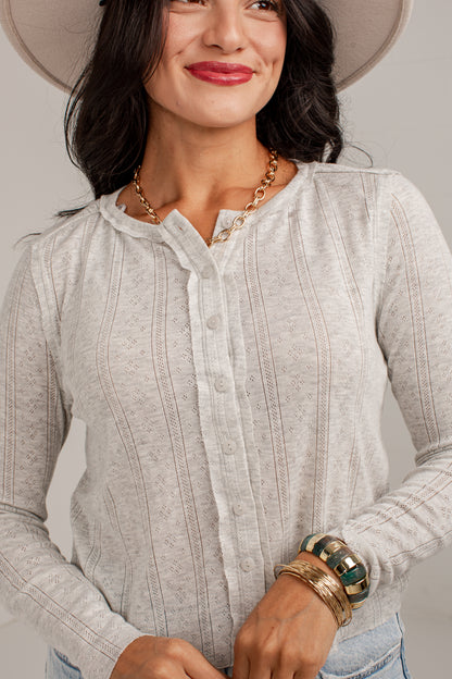 A woman wearing a light gray ribbed long-sleeve henley top with a button-down front, styled with a wide-brim hat, gold necklace, and stacked bracelets.