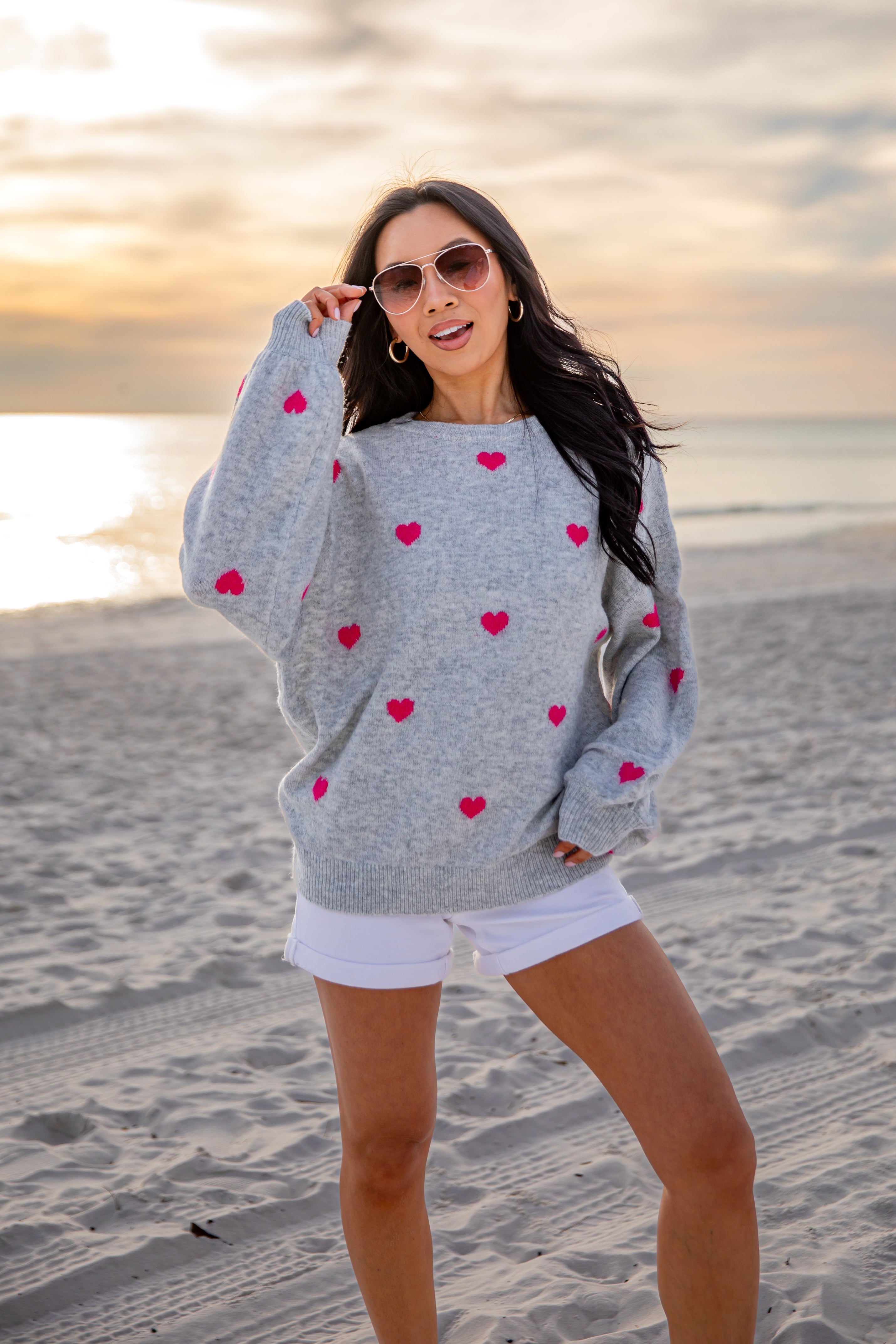 Woman wearing a gray sweater with red heart patterns on a beach at sunset.