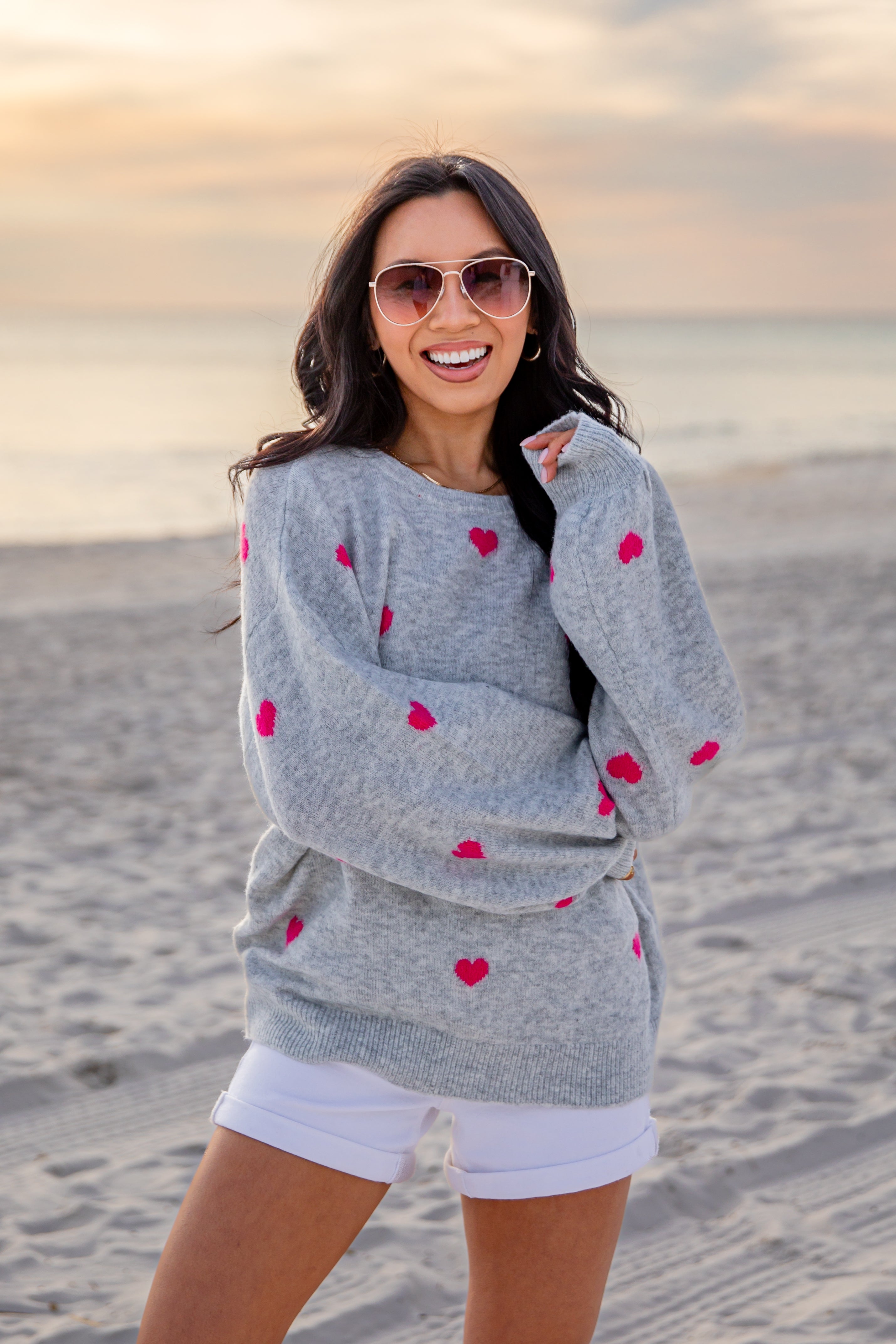 Woman wearing a gray sweater with red heart patterns on a beach at sunset.