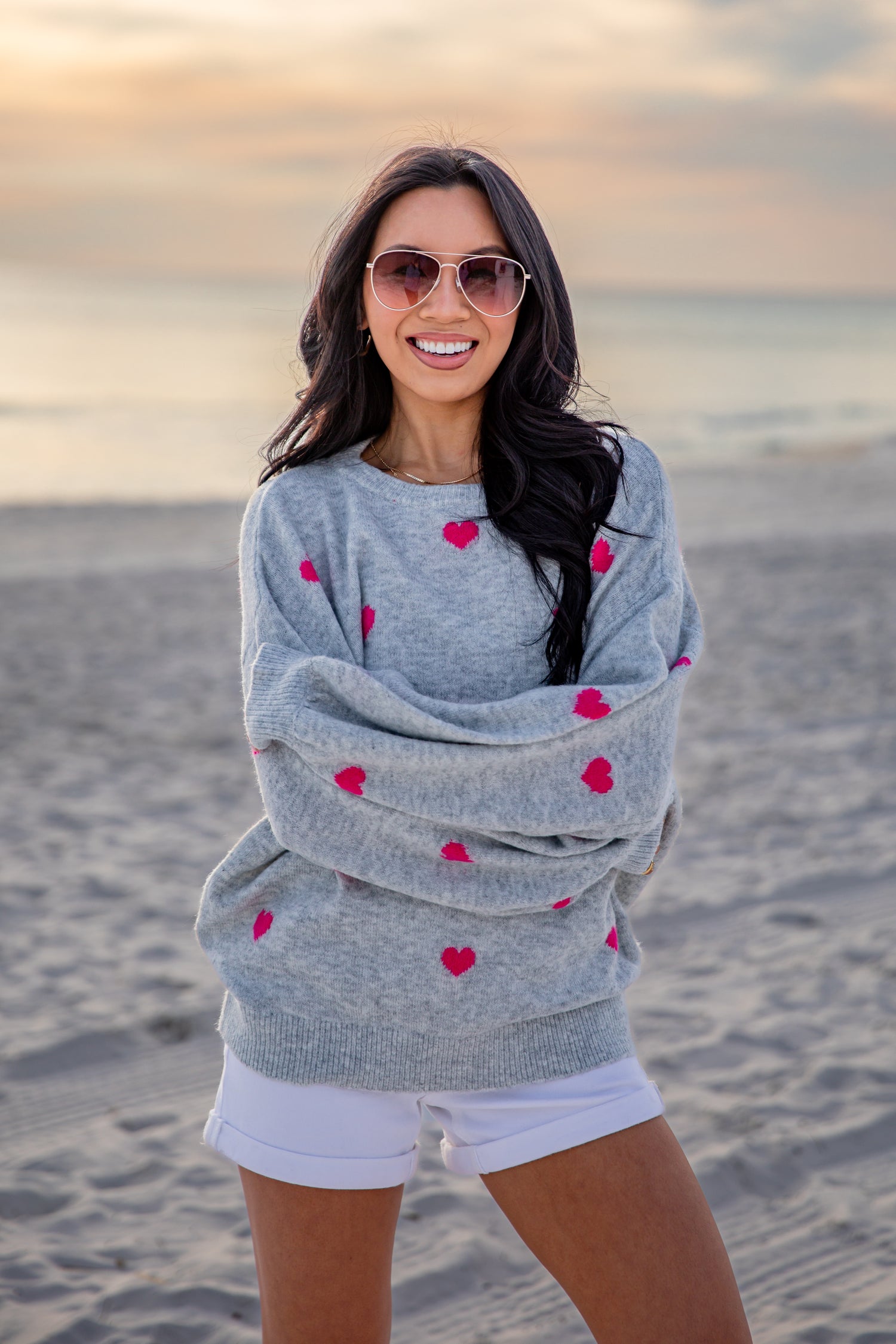 Woman wearing a gray sweater with red heart patterns on a beach at sunset.
