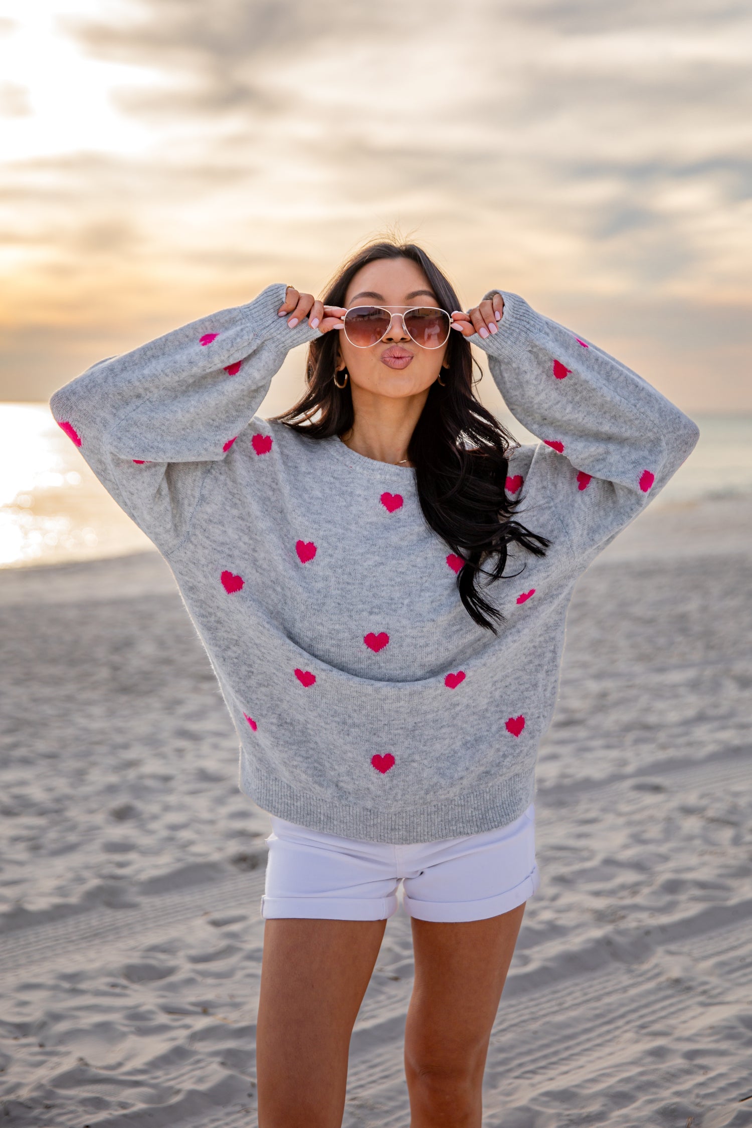Woman wearing a gray sweater with red heart patterns on a beach at sunset.