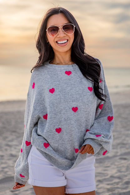 Woman wearing a gray sweater with red heart patterns on a beach at sunset.