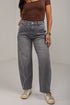 A model wearing a pair of wide-leg gray denim jeans, styled with a brown top and white sneakers, standing against a plain light gray wall.