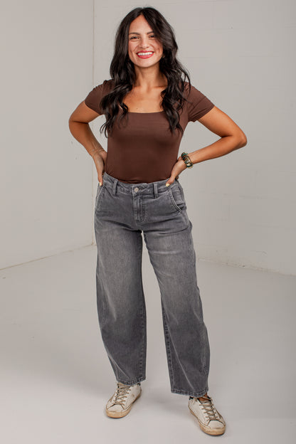 A woman modeling a brown scoop-neck short-sleeve top and gray wide-leg jeans, standing against a plain light gray studio background.