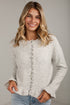 A smiling blonde woman wearing a light heather gray knit cardigan sweater with a button-down front and blue jeans, standing against a light gray wall.