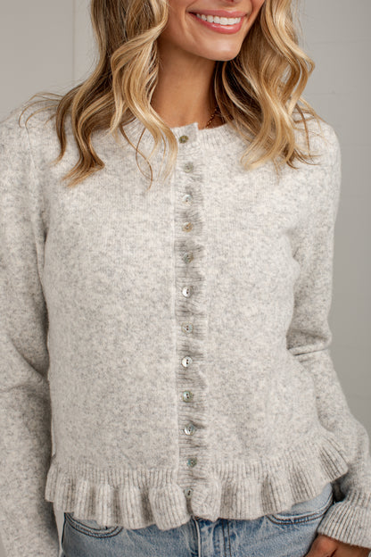 A woman wearing a light heather gray knit cardigan sweater with a button-down front and ruffled hem, paired with blue jeans.