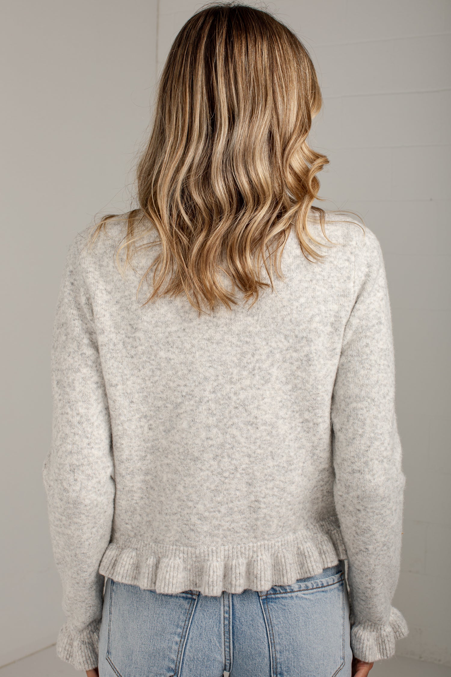 A woman viewed from behind wearing a light heather gray knit sweater with a ruffled hem and light blue jeans, set against a plain light gray wall.