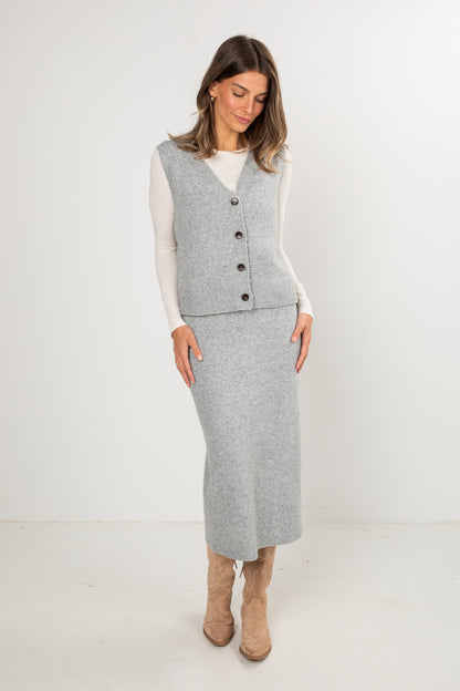 Woman wearing a grey knit midi skirt and grey buttoned sweater vest layered over a long-sleeve white top, styled with tan western boots, standing in a studio setting.
