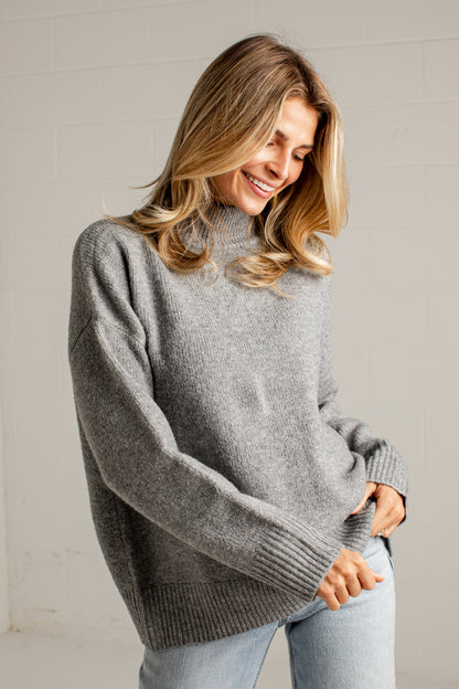 A woman wearing an oversized light gray knit sweater and light blue jeans, smiling and looking down in a bright indoor setting with a white brick wall background.