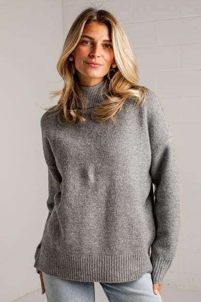 A woman wearing a heather gray ribbed knit mock neck sweater and light blue jeans, standing against a light gray brick wall.