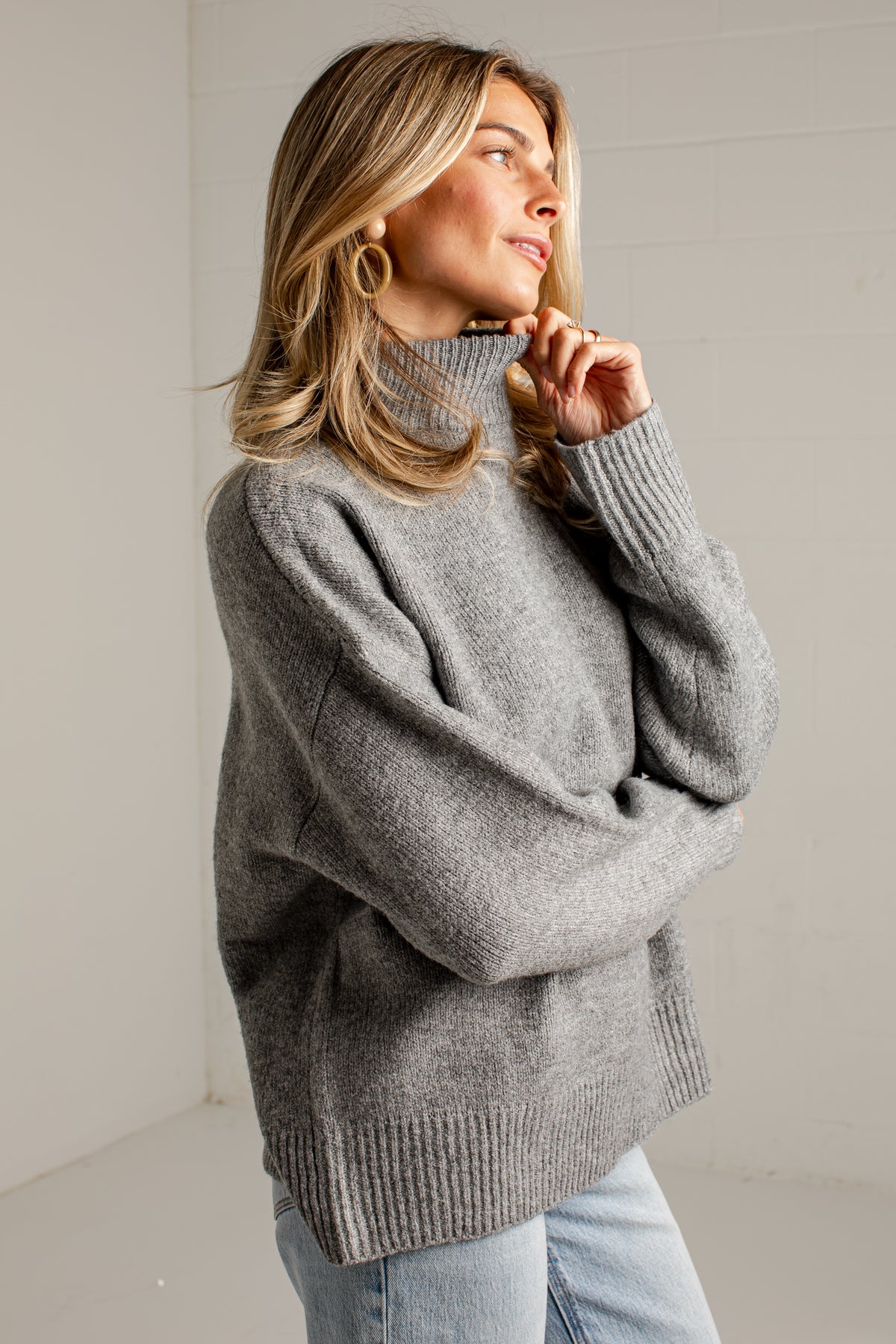 A woman wearing an oversized heather gray ribbed turtleneck sweater and light blue jeans, standing against a light gray wall.
