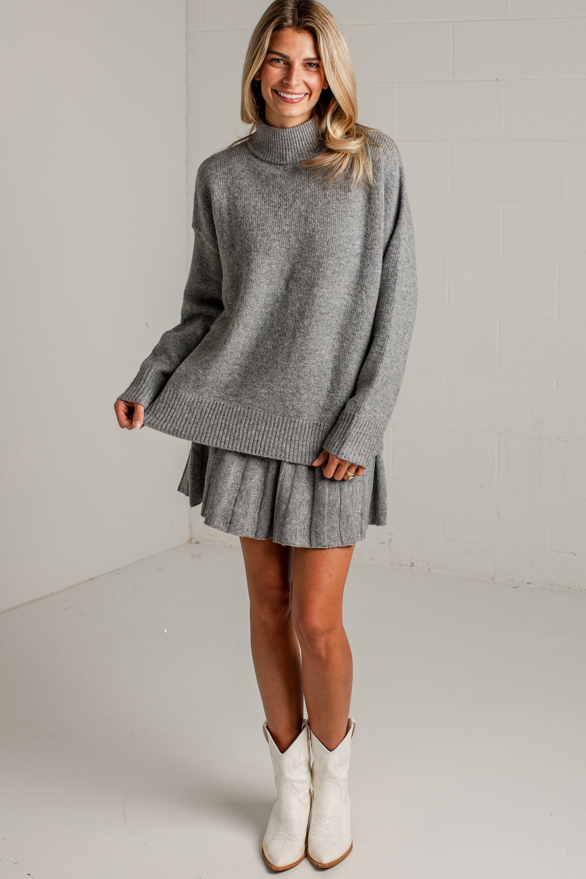 A woman modeling a gray knit turtleneck sweater dress with a pleated mini skirt, paired with white cowboy boots, in a bright studio setting.