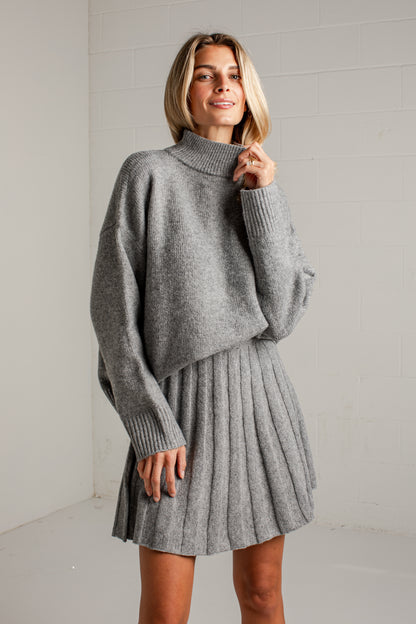 A woman modeling a gray knit mock-neck sweater and matching pleated mini skirt against a white brick wall background.