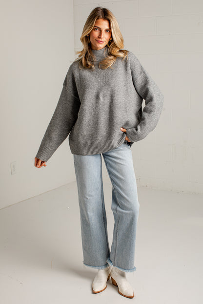 A woman modeling an oversized gray knit mock-neck sweater, paired with light-wash wide-leg jeans and white ankle boots, standing in a bright white studio.