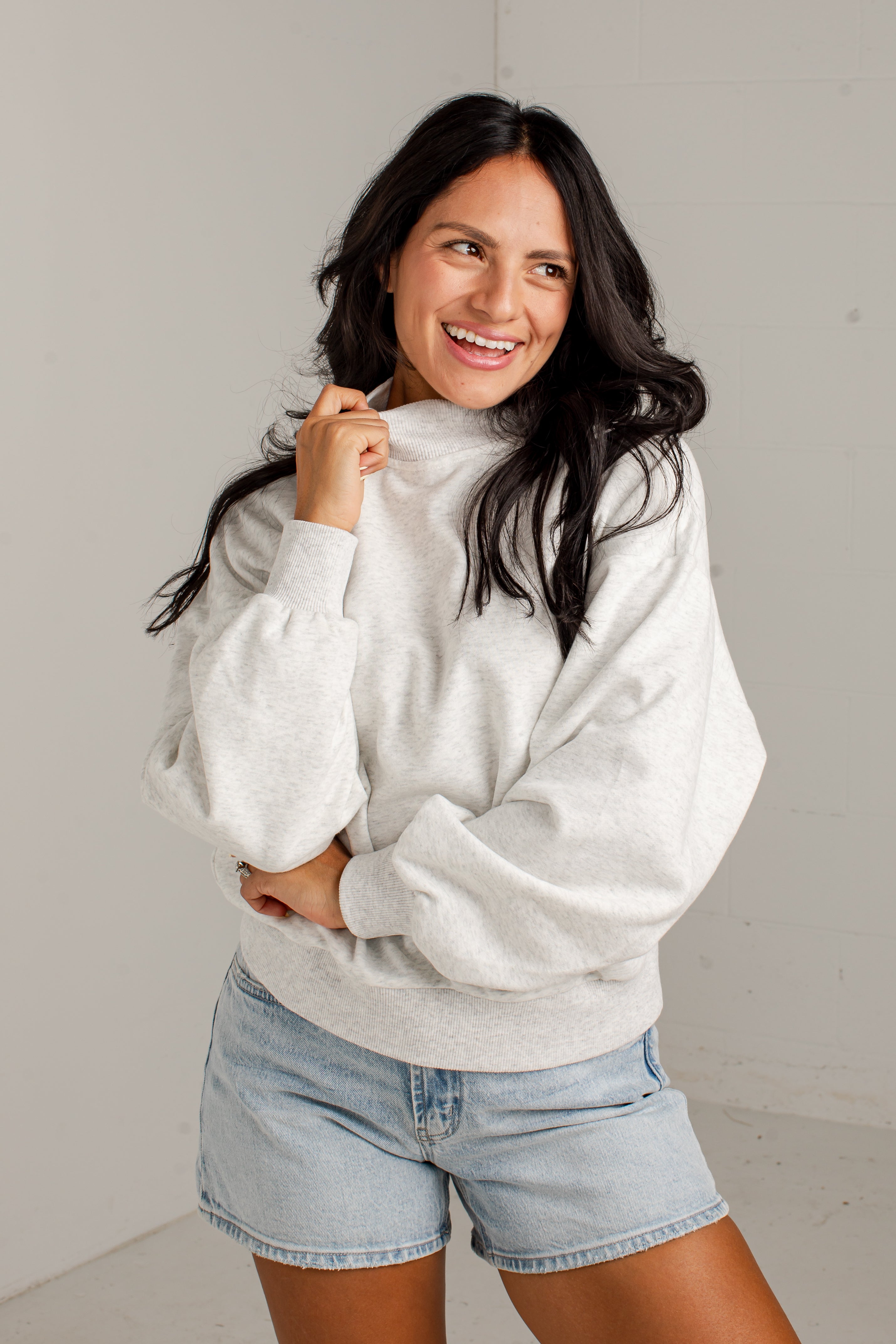A woman smiling while wearing a light gray oversized mockneck sweatshirt with balloon sleeves and light-wash denim shorts, standing against a plain light gray wall.