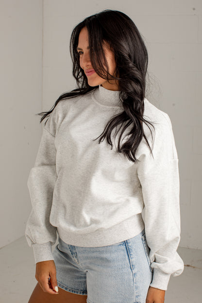 A woman wearing a light heather gray mock neck sweatshirt and light blue denim shorts, standing against a light gray wall.