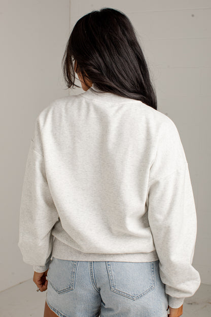 A person shown from behind wearing a light heather gray crewneck sweatshirt and light blue denim shorts against a plain light gray wall.