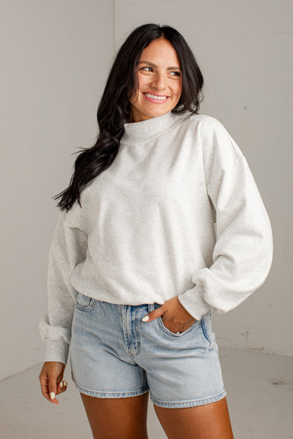 A woman wearing a light gray heathered mock neck sweatshirt and light-wash denim shorts, standing against a neutral light gray wall.