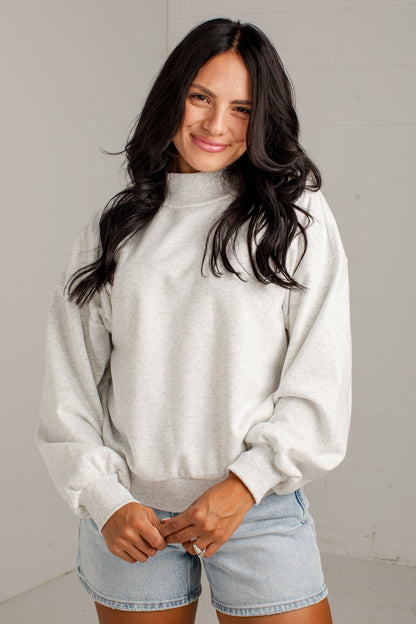A woman with long dark hair wearing a light heather gray mock neck sweatshirt and light blue denim shorts, standing against a light gray wall.