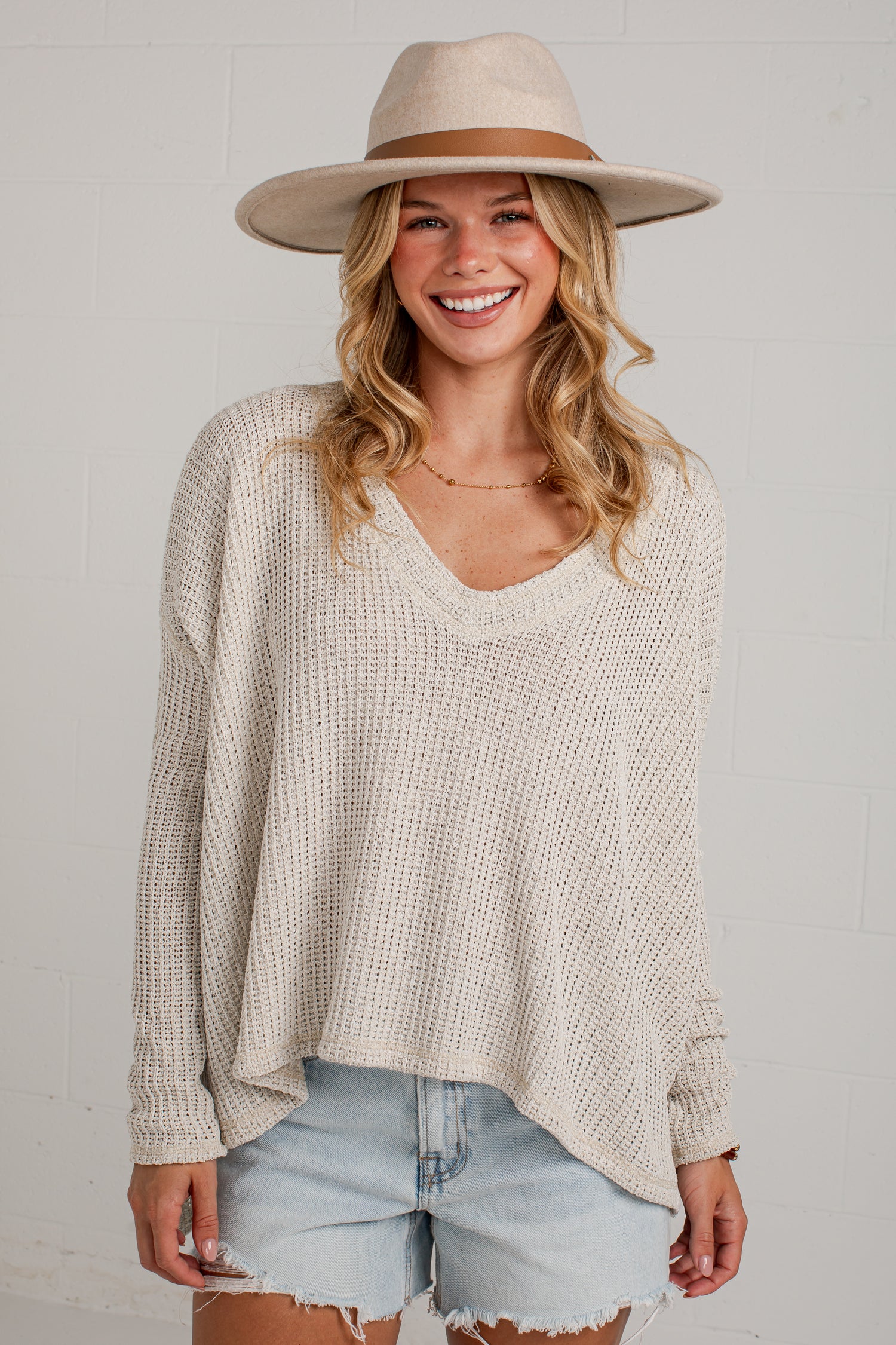 A woman wearing a cream-colored waffle-knit long-sleeve V-neck pullover sweater, light-wash denim shorts, and a beige wide-brim fedora hat, standing against a white brick wall.