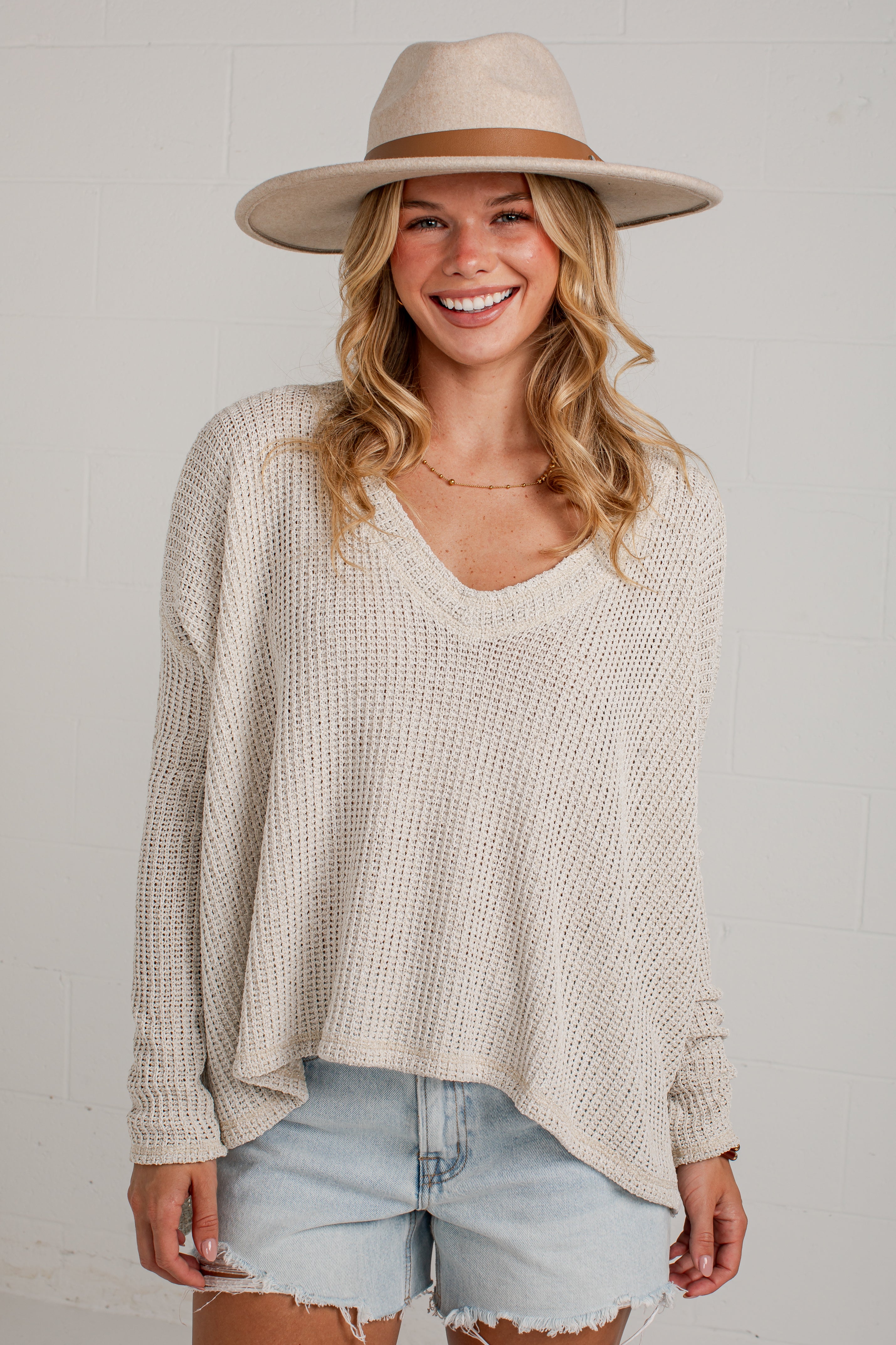 A woman wearing a cream-colored waffle-knit long-sleeve V-neck pullover sweater, light-wash denim shorts, and a beige wide-brim fedora hat, standing against a white brick wall.