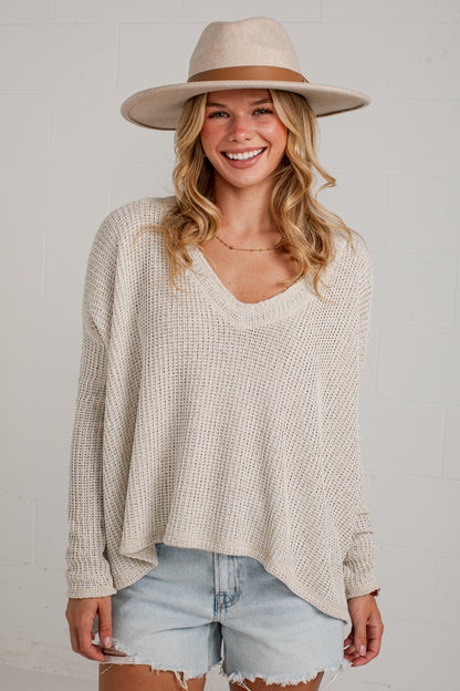 A woman wearing a cream-colored waffle-knit long-sleeve V-neck pullover sweater, light-wash denim shorts, and a beige wide-brim fedora hat, standing against a white brick wall.