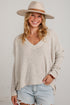 A woman wearing a beige waffle-knit V-neck sweater, a wide-brim beige hat, and light-wash denim shorts, standing against a white brick wall.